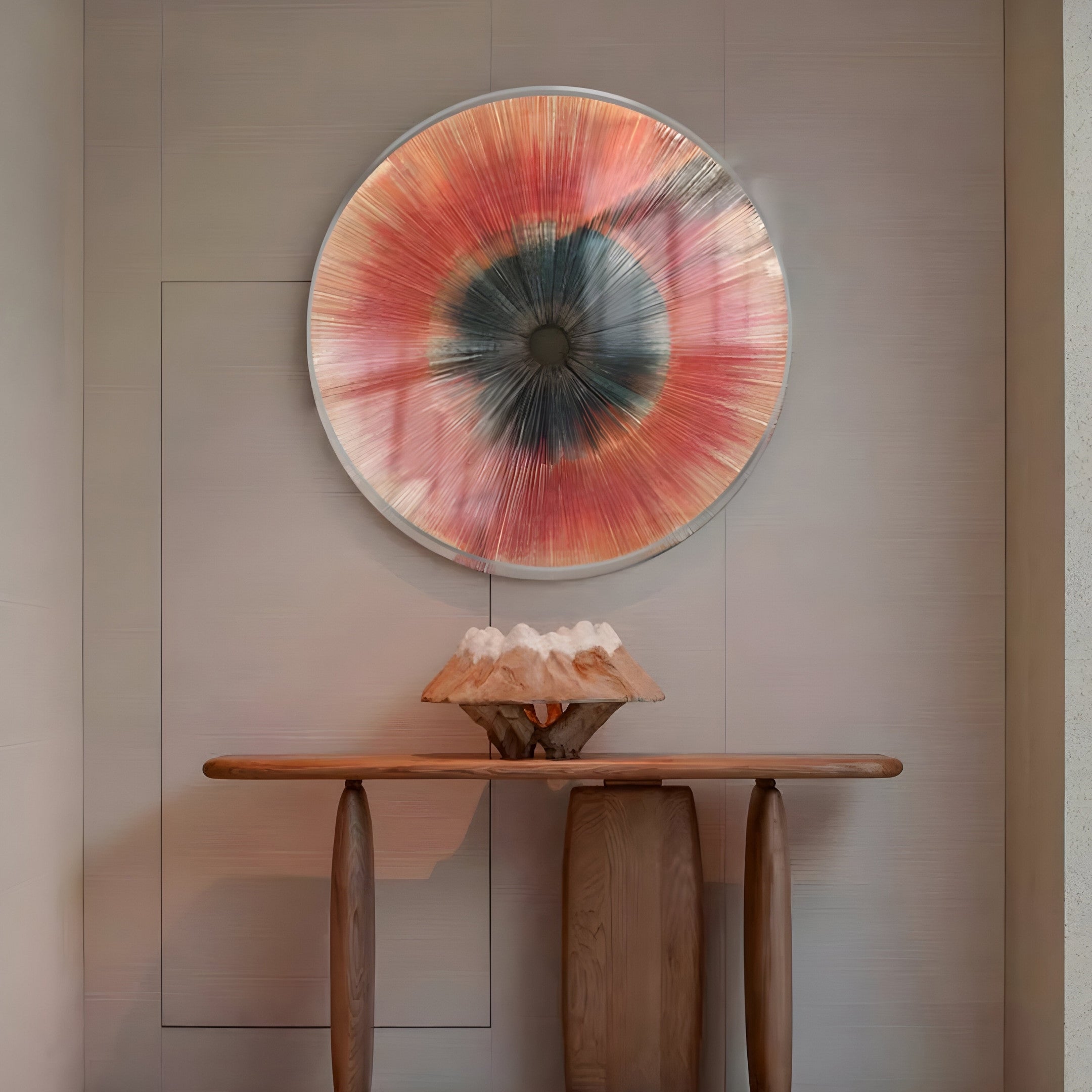 A wooden console table against a neutral wall displays a mountain-like décor. Above it, the Oculis Sunset Radial Circular Handmade 3D Paper Wall Art by Giant Sculptures hangs, featuring warm orange and pink hues with a dark center, epitomizing contemporary interiors with its handmade charm.