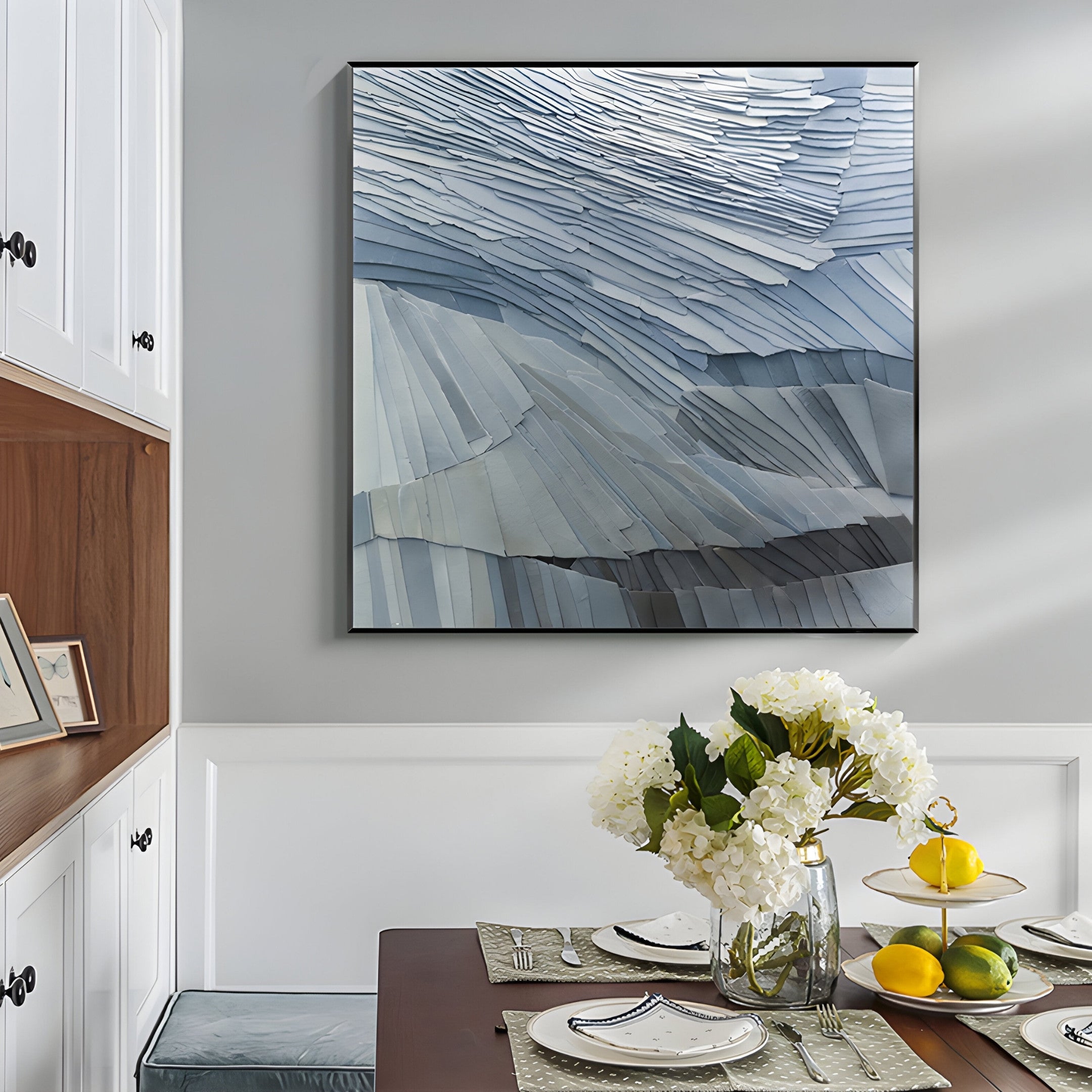 The dining area boasts a wooden table set for four, adorned with a glass vase of white flowers and a bowl of lemons. Above, the Terrane Tranquil Blue Layered Abstract Wall Art by Giant Sculptures enhances the wabi-sabi style of white cabinets with black handles on the left.