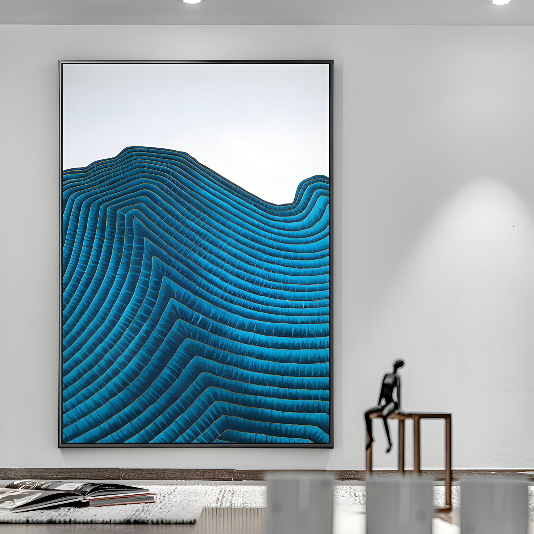 A modern luxury decor interior showcases the Ripplewave II Abstract Blue Decorative Enamel Wall Art by Giant Sculptures, highlighting blue wavy lines reminiscent of mountains. A small cloisonné enamel figurine adorns a minimalist table, softly illuminated in the foreground.