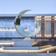 The Crescent Abstract Steel Outdoor Sculpture (120cm) by Giant Sculptures features a silver crescent moon on a geometric pedestal with reflective patterns, enhancing any modern garden with its serene and contemporary design, perfect against a building facade with vertical light strips.