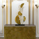 The Celest Abstract Steel Sculpture by Giant Sculptures, a 93cm gold art piece with swirling designs on a black base, is displayed atop a gold-patterned cabinet with intricate spiral motifs. Ideal for modern spaces, its set against an elegant white wall with gold accents and two decorative sconces.