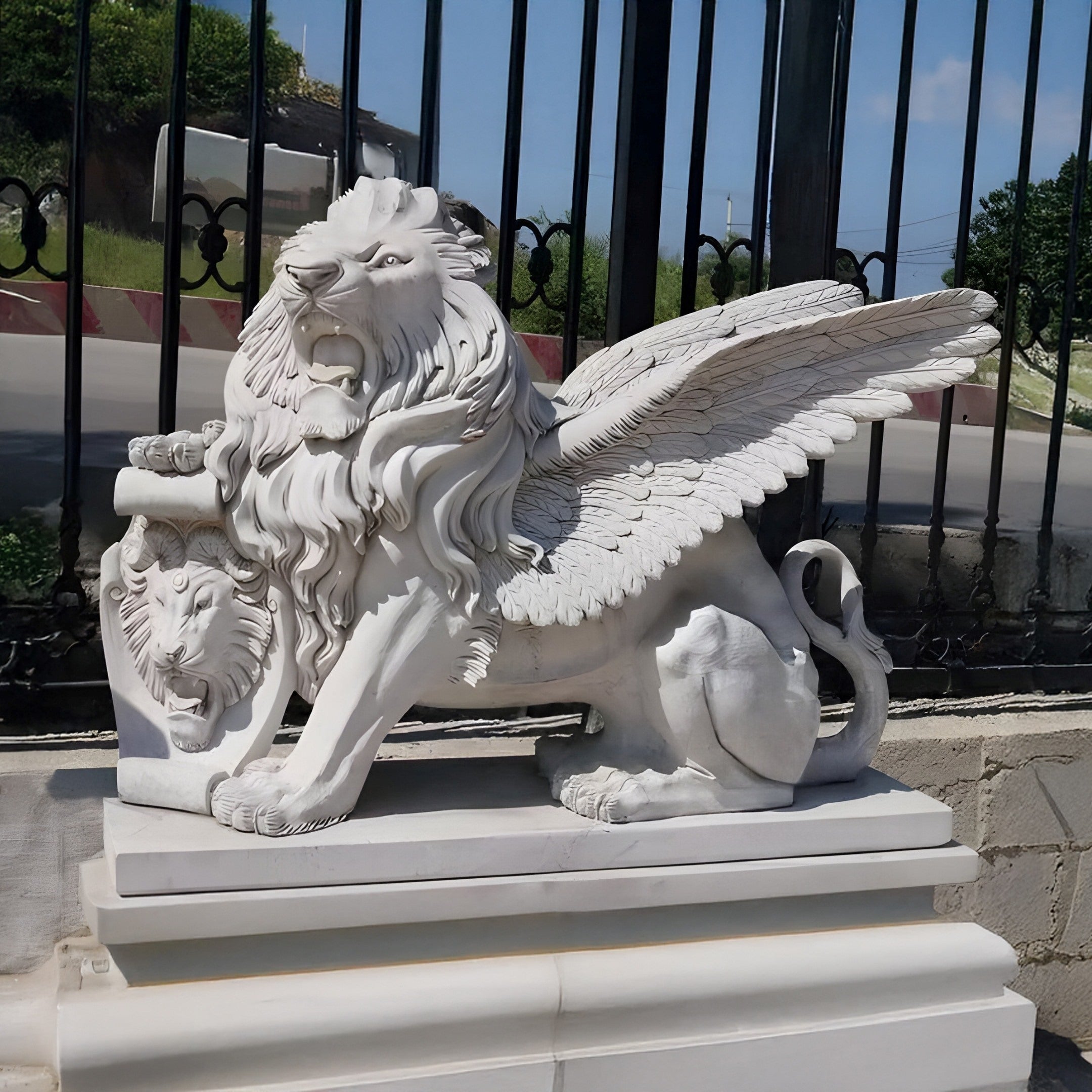 The Giant Sculptures Giant Winged Lion Marble Outdoor Sculpture, customized with a lions head emblem on a scroll, sits majestically on a pedestal before an iron fence. It enhances the garden décor amid lush greenery and clear blue skies.