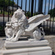 The Giant Sculptures Giant Winged Lion Marble Outdoor Sculpture, customized with a lions head emblem on a scroll, sits majestically on a pedestal before an iron fence. It enhances the garden décor amid lush greenery and clear blue skies.