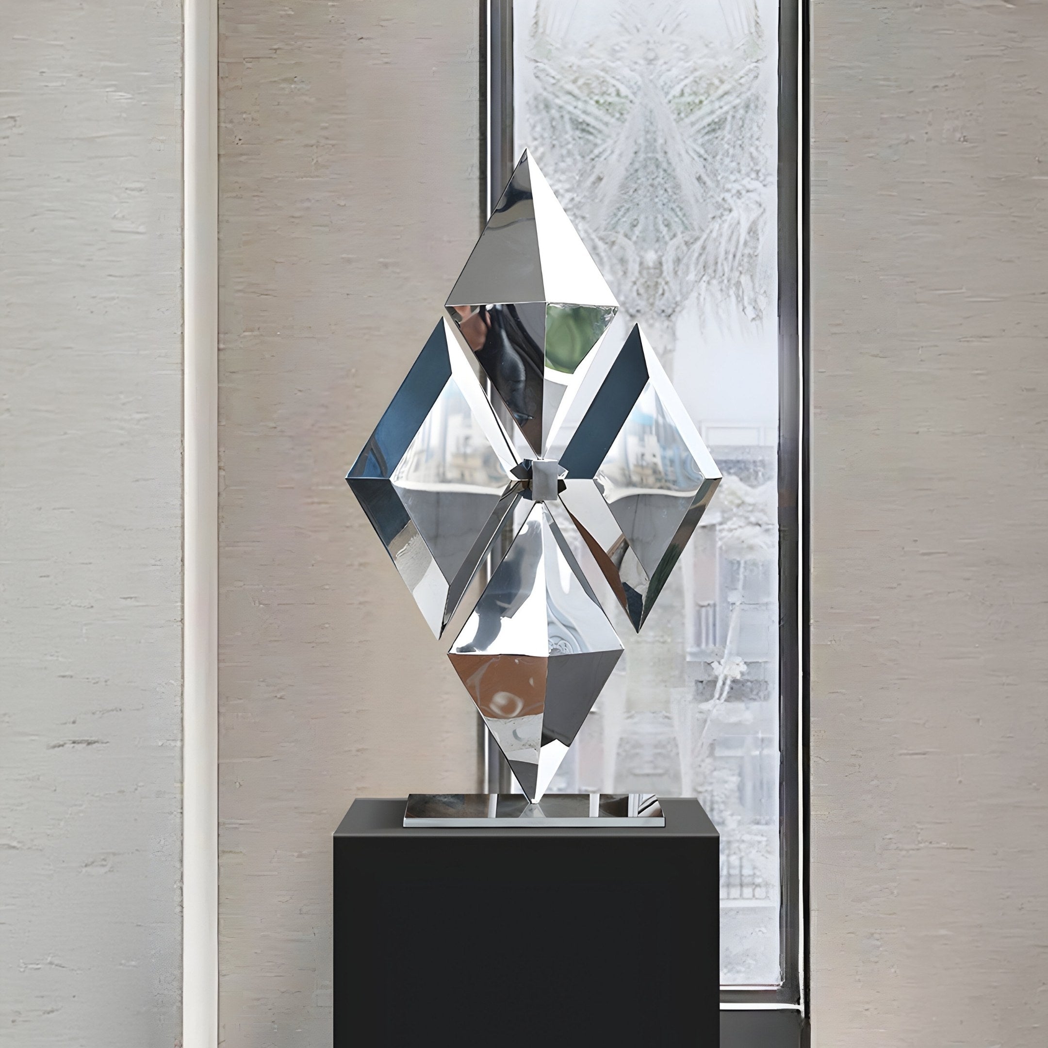 The Diamond Geometric Steel Sculpture - 103cm by Giant Sculptures is elegantly showcased on a black pedestal, its diamond-like facets beautifully distorting and reflecting ambient light and surroundings in front of a large window.