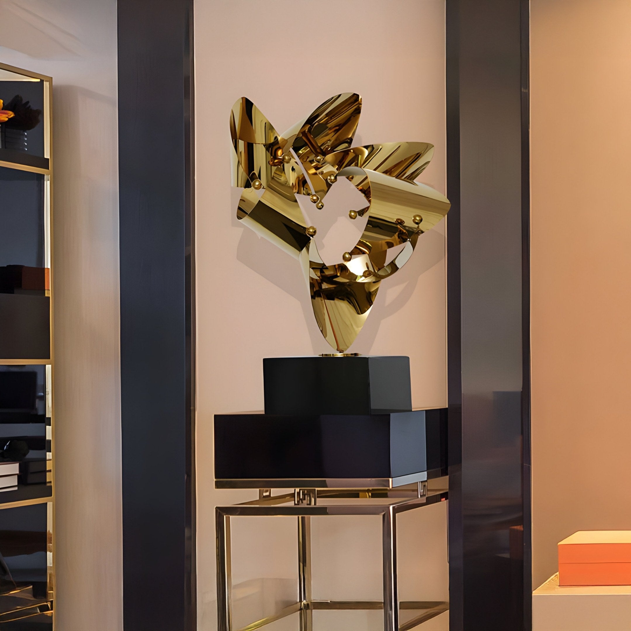 The Crest Gold Abstract Steel Sculpture by Giant Sculptures, featuring mirrored surfaces and angular shapes, is showcased on a black pedestal against a neutral wall. Nearby, shelves and decor complete the modern display.