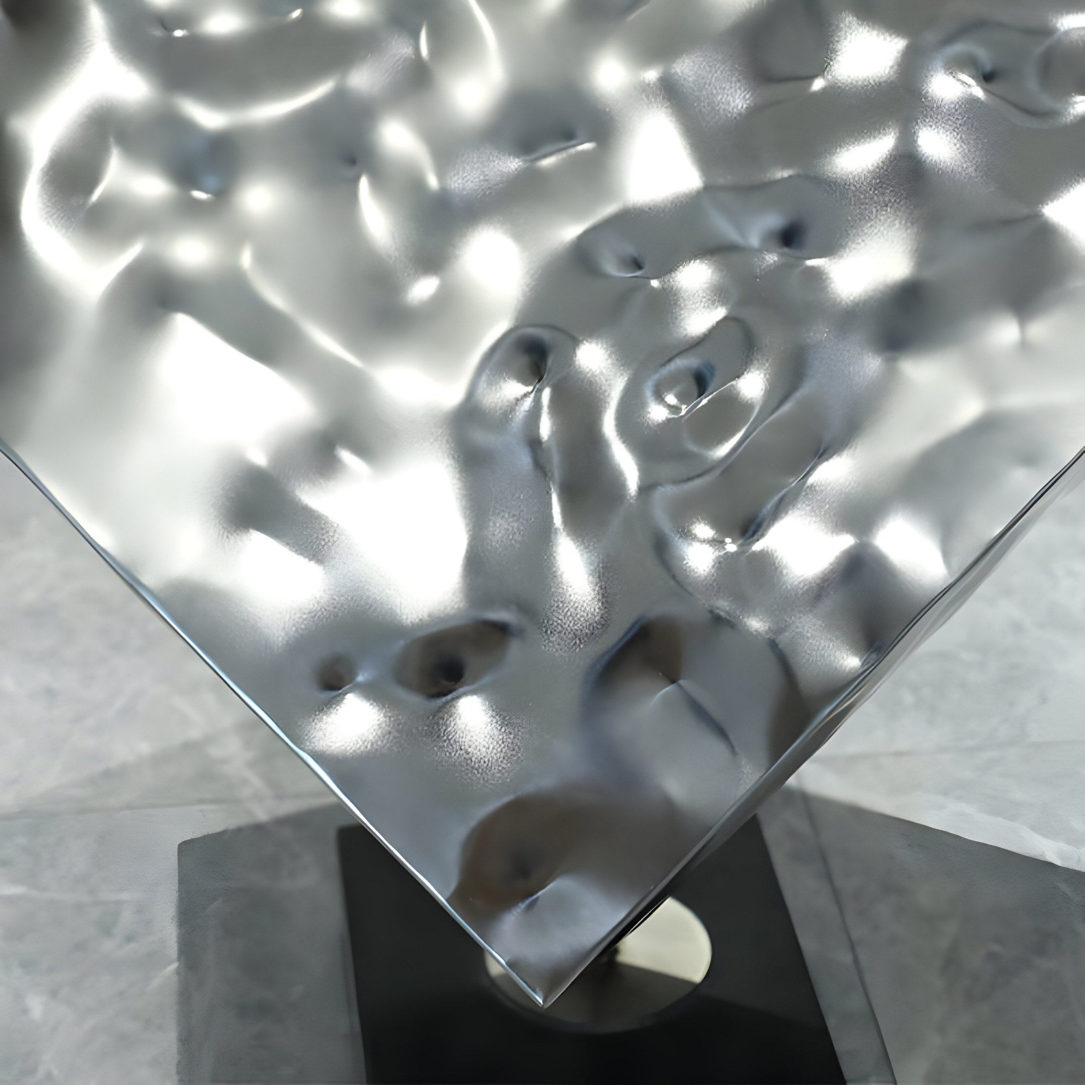 The Cubic Geometric Steel Sculpture - 75cm by Giant Sculptures features a stainless steel, textured surface that gracefully rests on a black base. Its reflective design creates fluidity with light and shadow playing over its form, complemented by the visibility of the marble floor below.