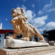 The Giant Sculptures Giant Lion Yellow Marble Outdoor Sculpture, standing 240cm tall, is intricately carved and polished. Under a blue sky with clouds, it majestically rests on a matching base with buildings in the background and is customizable for outdoor spaces.