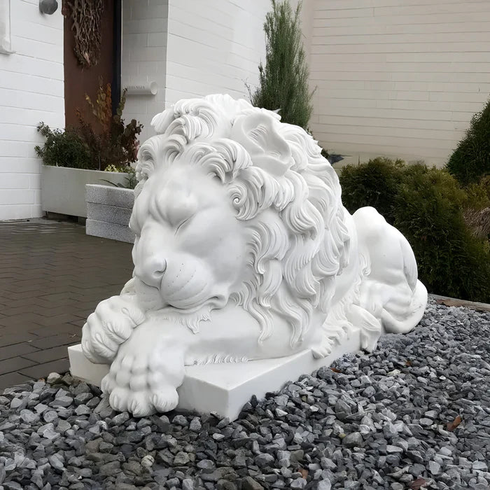 The Giant Sleeping Lion Marble Outdoor Sculpture by Giant Sculptures showcases a white marble lion with detailed mane and paws, lying on gray stones among greenery. The closed eyes add elegance, set against a backdrop of a white building wall and plants, exemplifying exquisite garden decor.