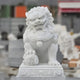 A hand-carved marble sculpture, the Chinese Guardian Lion by Giant Sculptures, is intricately detailed and stands proudly on a pedestal. With a fierce expression and decorative features, including a ball beneath its paw, this magnificent piece displays the artistry of ancient design against blurred stone structures in the background.