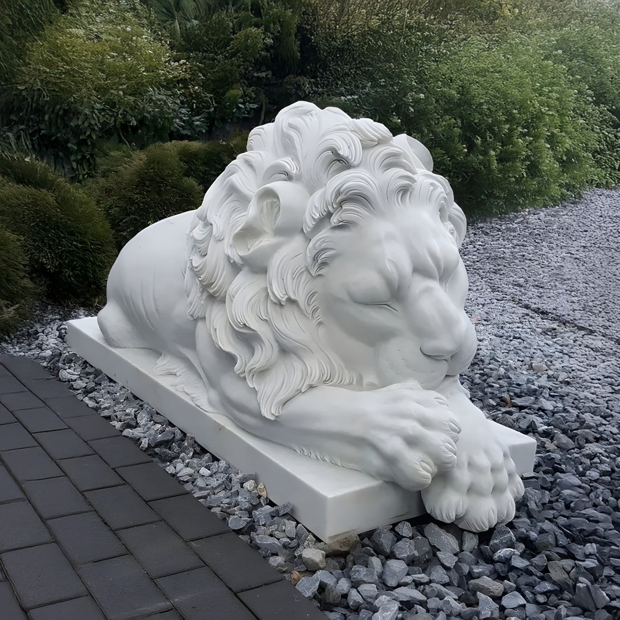 The Giant Sleeping Lion Marble Outdoor Sculpture - Customised by Giant Sculptures serves as a stunning piece of garden decor. Crafted in large, white marble, it rests gracefully on a bed of gray pebbles with eyes closed. The intricately detailed mane and relaxed pose create a striking contrast with the surrounding greenery and paved walkway, making it a standout outdoor sculpture.