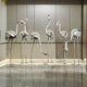 Flamingo Stainless Steel Outdoor Sculptures by Giant Sculptures, available in 94/147cm sizes, are elegantly arranged on a reflective marble floor in a modern minimalist room. The various poses of these sculptures form an artistic display against a neutral-colored wall.