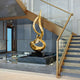 A 173cm Flare Gold Abstract Steel Sculpture by Giant Sculptures sits on a black pedestal at the base of a staircase in a luxurious, contemporary interior with glass walls, marble flooring, and sleek minimalist design.