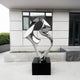 The Flow III Silver Organic Steel Sculpture (96cm) by Giant Sculptures, with its shiny abstract design, rests on a black base before a modern building featuring large windows and grey panels. Its weather-resistant construction complements the surrounding small greenery in planters.