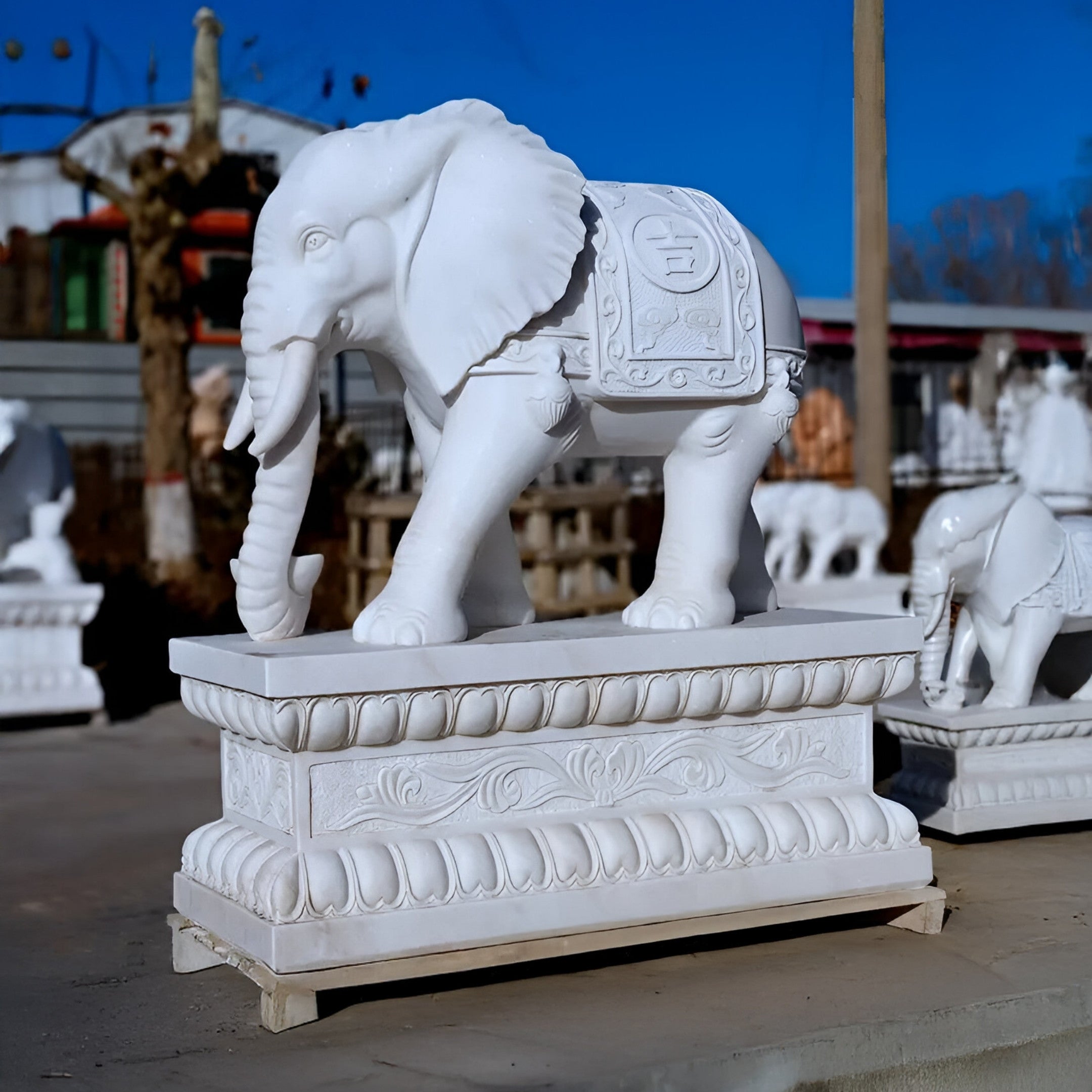 The Lucky Chinese Elephant Marble Outdoor Sculpture by Giant Sculptures stands on a decorative pedestal outdoors, showcasing intricate carvings on its body and trunk, with other stone statues fading into the clear blue sky, portraying timeless artistry.