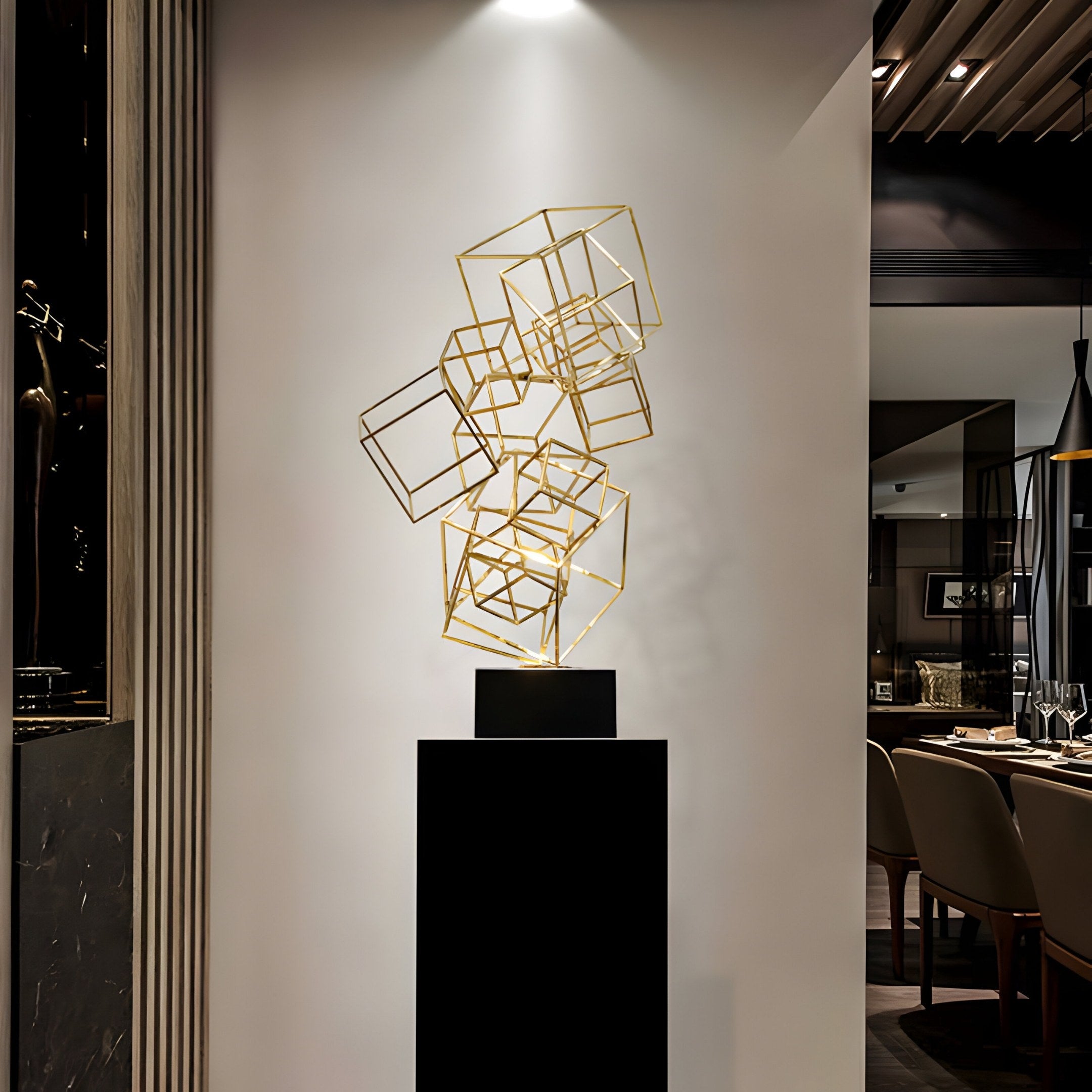 The Frame Shift Geometric Steel Sculpture by Giant Sculptures, featuring interconnected stainless steel cubes with a gold finish, stands on a black pedestal against a white wall. It enhances the modern space with dining tables and ambient lighting, adding an artistic flair to the setting.