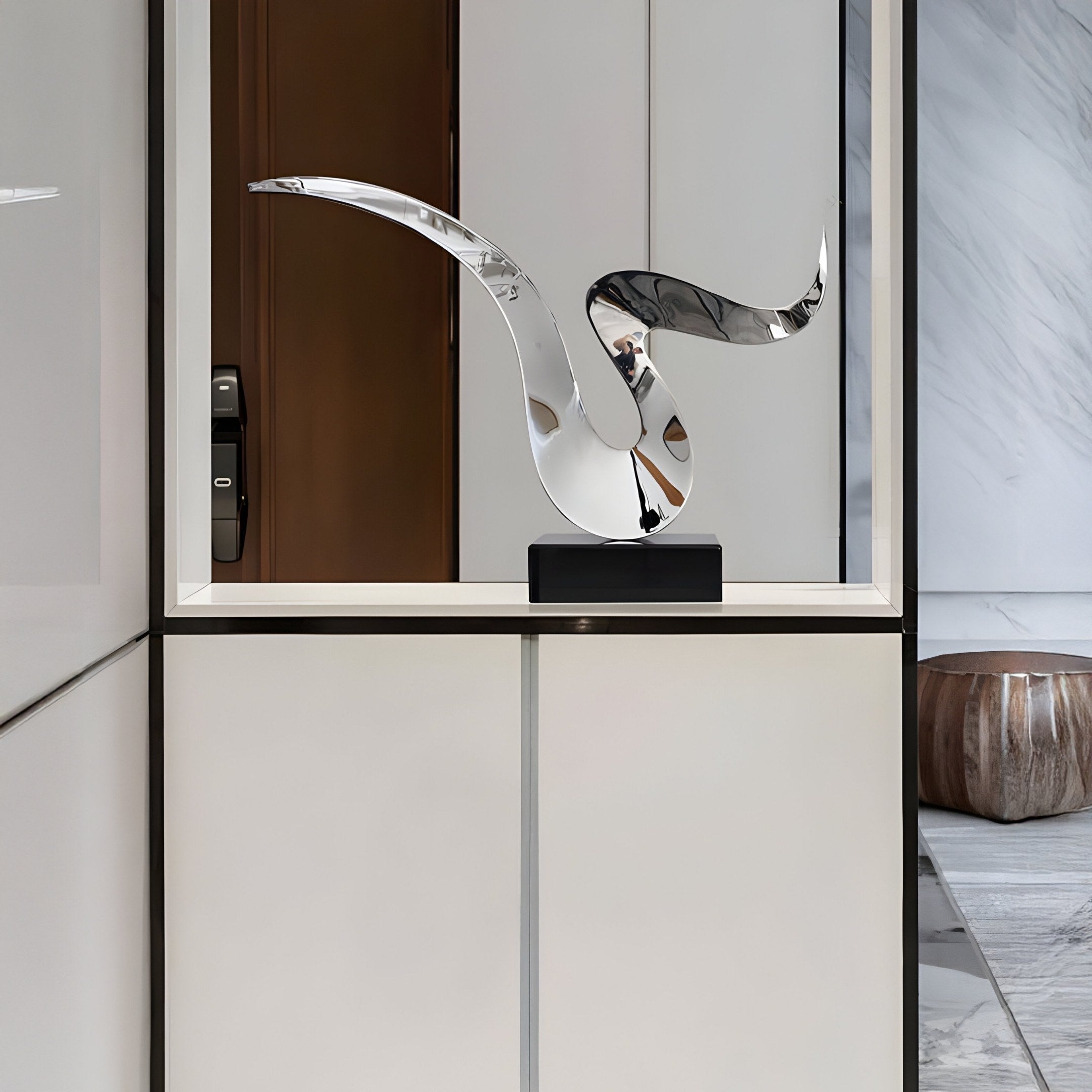 A sleek modern room features the Whisper Silver Abstract Steel Sculpture by Giant Sculptures on a white pedestal, reflecting its surroundings. The space has minimalist white walls and a marble floor, with a brown leather pouf in the corner enhancing the contemporary aesthetic.