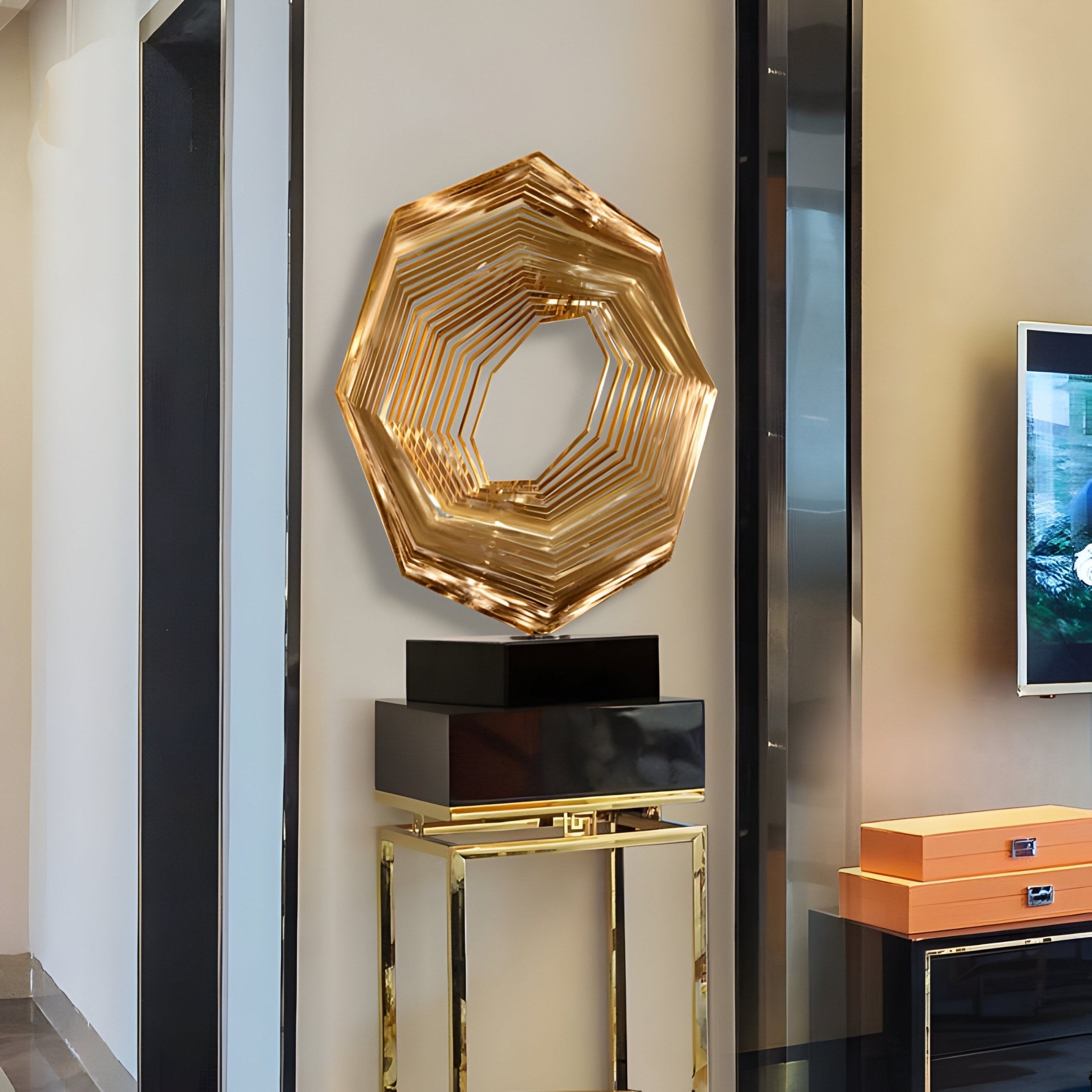 The Hexara Gold Geometric Steel Sculpture by Giant Sculptures, a 70cm abstract composition of angular shapes in a circle, hangs above a glossy black and gold stand. Orange boxes are nearby with the TV screen visible to the right.