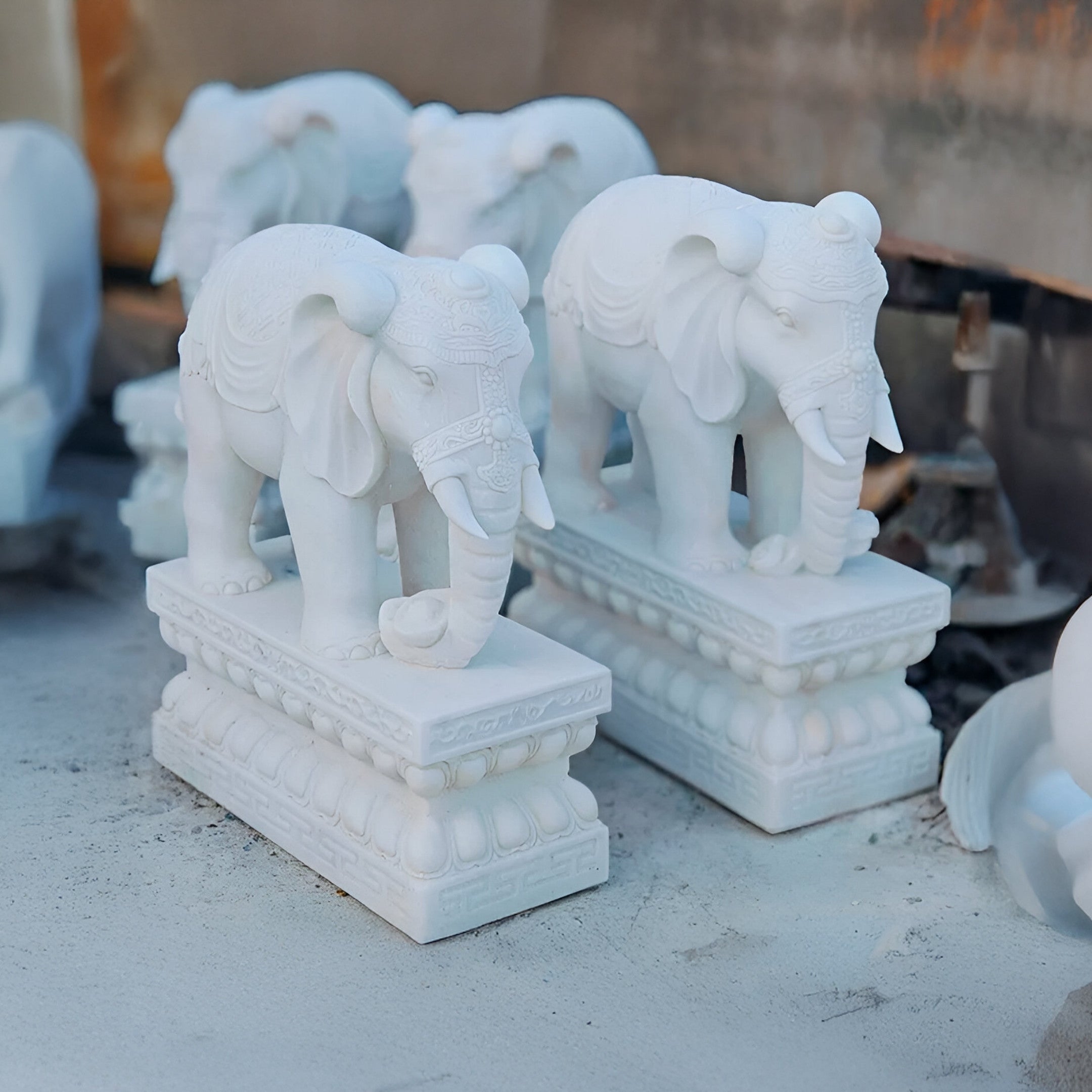 The Indian Elephant Marble Outdoor Sculpture - 120cm by Giant Sculptures features statues with intricate carvings on rectangular bases, placed elegantly among other captivating pieces.