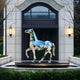 The Horse II Geometric Steel Outdoor Sculpture by Giant Sculptures, standing 182cm tall on a round pedestal, graces an elegant building entrance. Surrounded by trimmed hedges and bare branches, its flanked by windows and warm-lit lanterns in this modern outdoor setting.