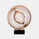 Circular copper sculpture with concentric rings creating an illusion of motion, mounted on a black base.