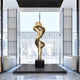 The Liquid Aura Gold Steel Sculpture by Giant Sculptures, standing elegantly at 238cm on a black pedestal, gleams in natural light through large windows in a bright room with sleek white walls and comfy seating.