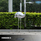 The Crane Contemporary Outdoor Steel Sculpture (98/158cm) by Giant Sculptures features a chrome-finished crane with long legs and a pointed beak. It stands on concrete, reflecting its surroundings, with green bushes in the background. The text VERSION 2 is in the bottom left corner.