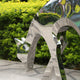 Close-up of a stainless steel deer sculpture with angled legs and reflective surfaces in a landscaped garden.