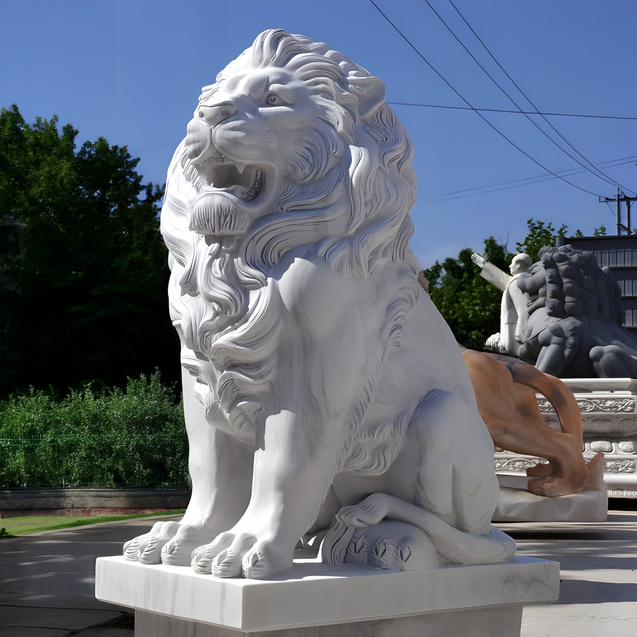 The Giant Lion Marble Outdoor Sculpture - Customised by Giant Sculptures majestically sits on a pedestal outdoors, facing forward with its full mane and open mouth. In the background, trees frame another sculpture partially visible. The clear blue sky enhances this awe-inspiring outdoor piece.