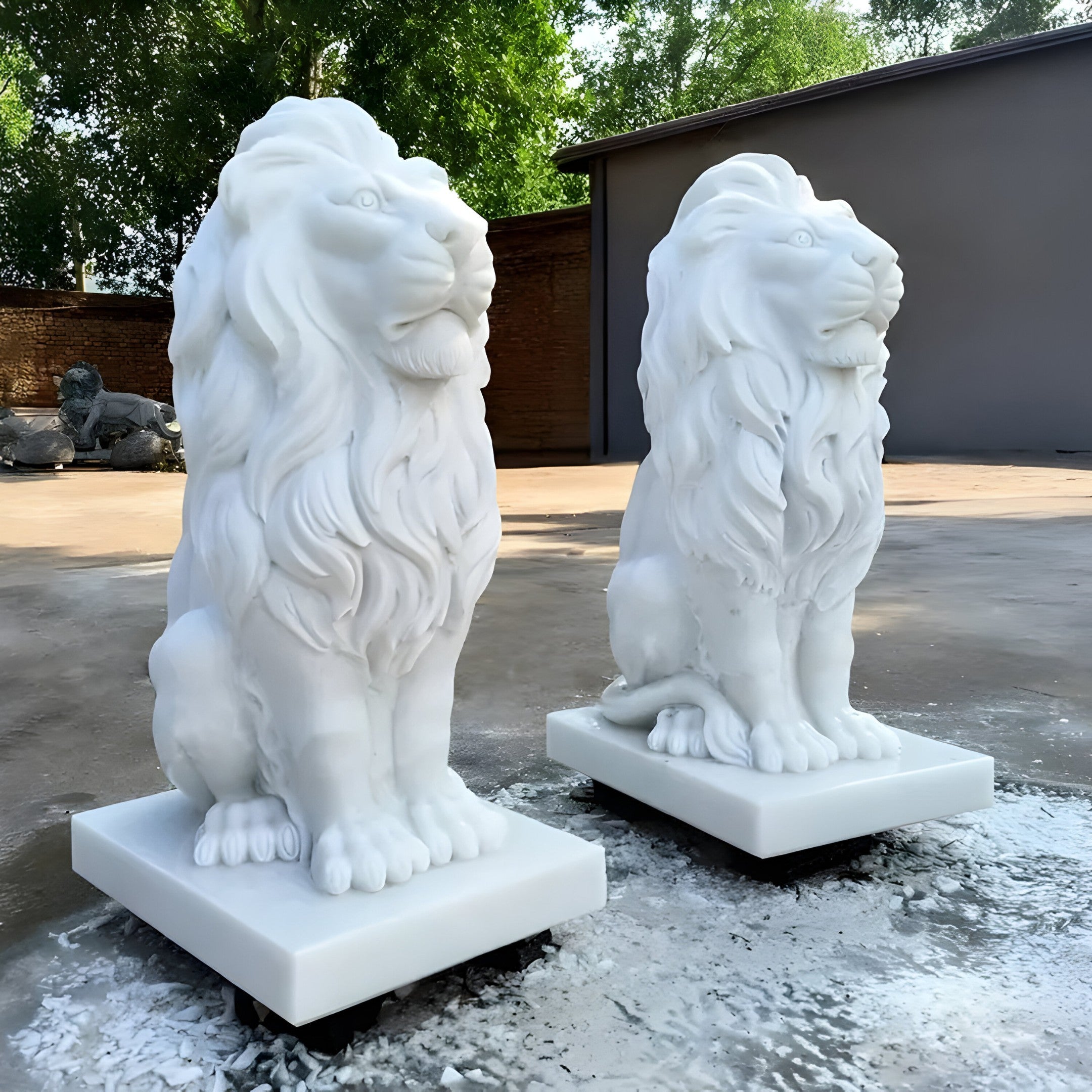 The "Giant Gracious Lion Marble Outdoor Sculpture - Customised" by Giant Sculptures features two handcrafted marble lion statues majestically positioned on square bases in an outdoor setting. These detailed sculptures capture the lions in a seated pose, radiating elegance and strength. This opulent garden art piece boasts a backdrop of trees and a building visible in the background.