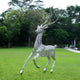 The Arcane Stag Steel Outdoor Sculpture by Giant Sculptures, standing at 205cm, is crafted in stainless steel with intricate wire patterns. Its elongated, abstract antlers create a whimsical feel amidst the grassy field and trees, with pathways and seated park-goers enhancing the serene atmosphere.