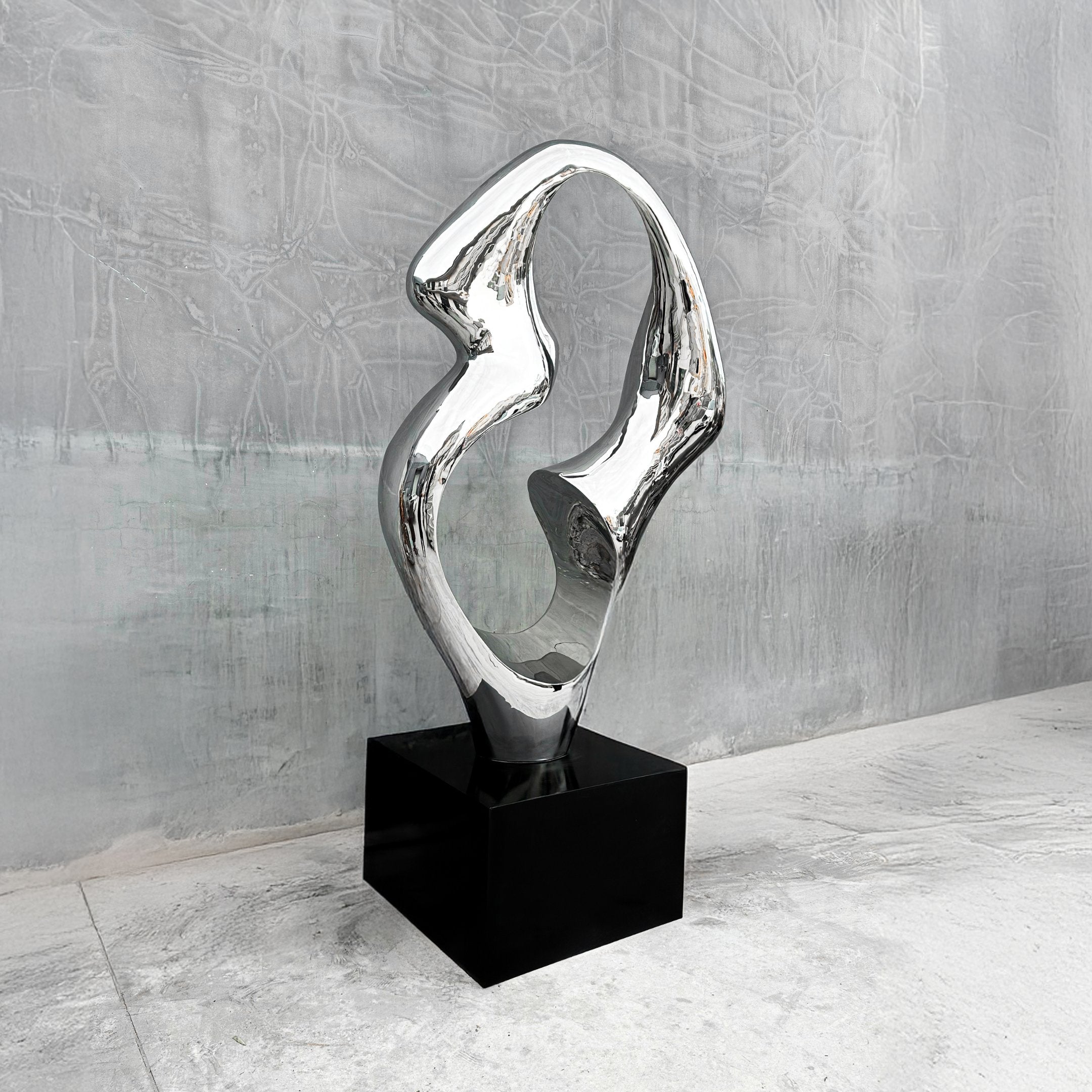 The Flow III Silver Organic Steel Sculpture by Giant Sculptures, featuring fluid, curving shapes, serves as a versatile centerpiece on a black square pedestal. It gleams against the textured gray background atop the light stone floor.