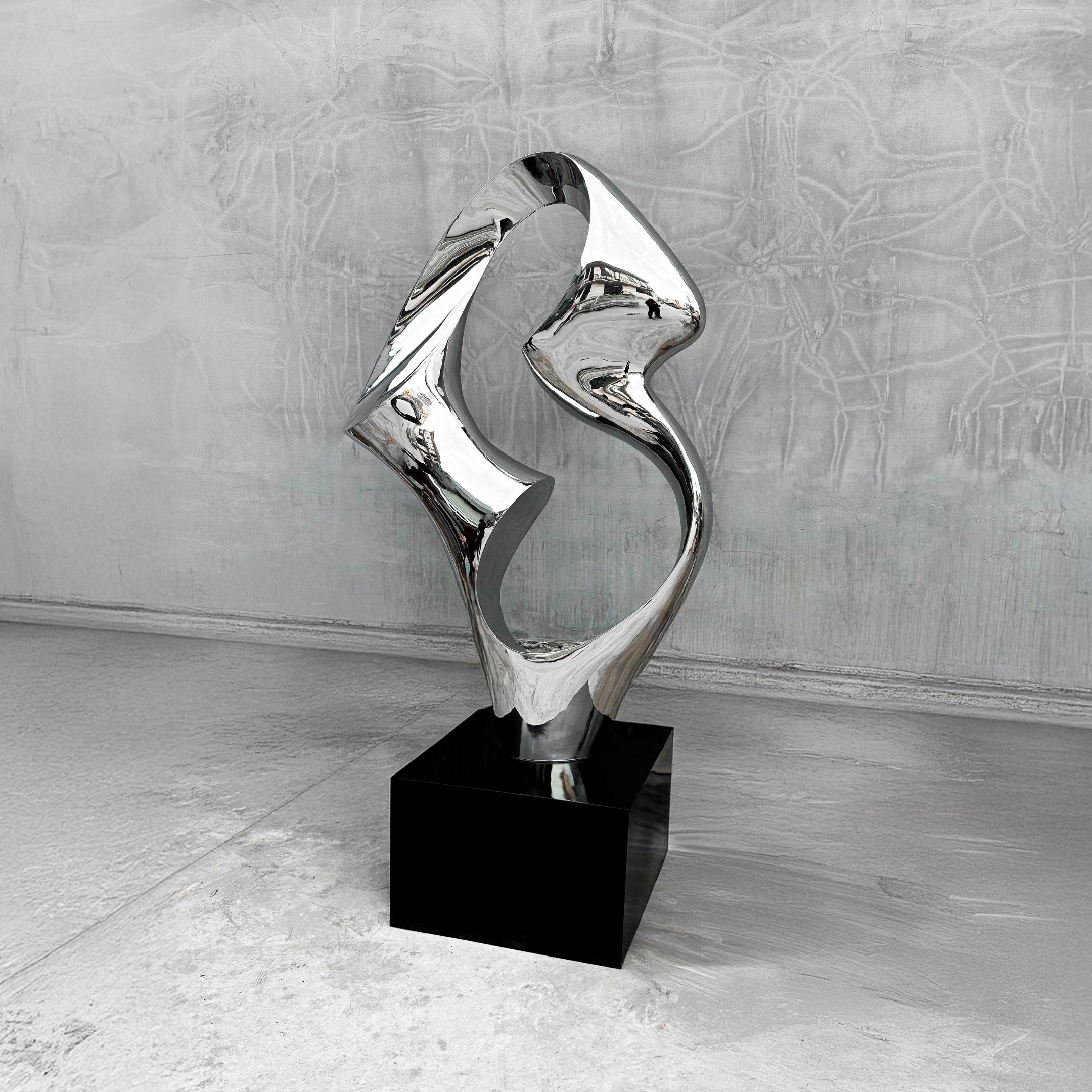 The Flow III Silver Organic Steel Sculpture by Giant Sculptures, standing at 96cm, features a modern abstract design with smooth, curving lines of silver organic steel on a black pedestal. Its weather-resistant build reflects light dynamically against a textured gray wall.