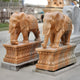 Two giant yellow marble elephant sculptures, intricately hand-carved and adorned with ornate designs stand outdoors under a partly cloudy sky. The primary focus is on the exquisite Elephant Yellow Marble Outdoor Sculpture - 180cm by Giant Sculptures, with a third blurred statue in the background.