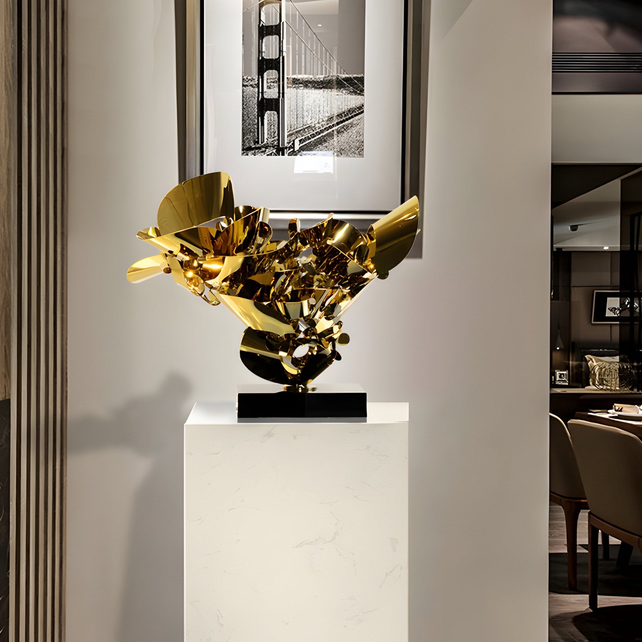 The Petal Fusion Gold Organic Steel Sculpture by Giant Sculptures, standing 70cm tall, adorns a white pedestal in a modern space. Above is a black-and-white photo of a suspension bridge, enhancing the elegant scene with tables and chairs awaiting in the background.