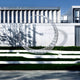 The Crescent Wireframe Outdoor Steel Sculpture by Giant Sculptures, standing 242cm tall, graces the lawn before a modern building with a grid-patterned facade. Shadows on the white walls enhance its beauty under a clear blue sky.