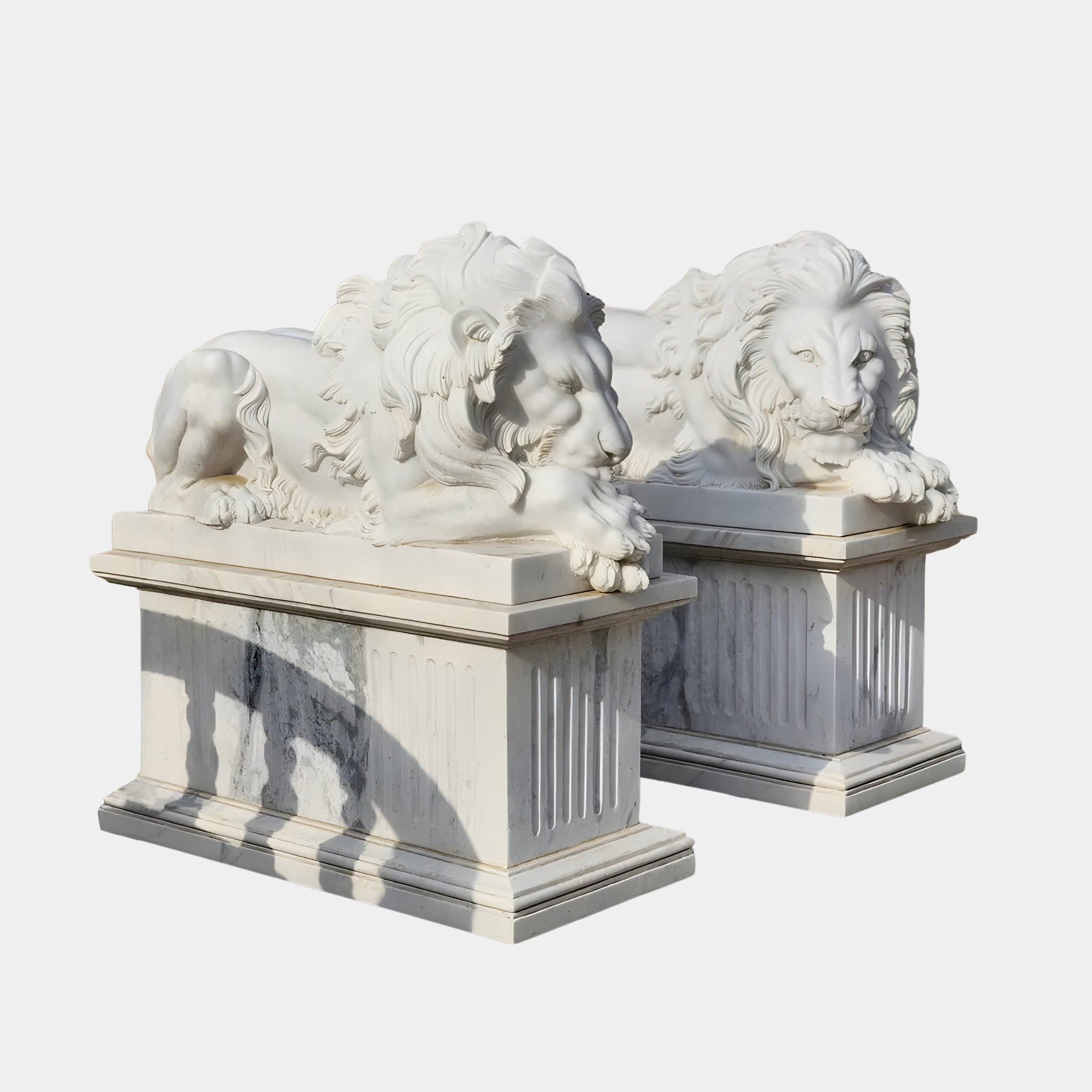 The Giant Lying Lion Marble Outdoor Sculptures from Giant Sculptures rest on rectangular pedestals. Featuring intricately detailed manes, the lions are positioned side by side. Bright lighting casts shadows on the pedestals, enhancing their presence against a plain background and emphasizing these sophisticated garden decor pieces.