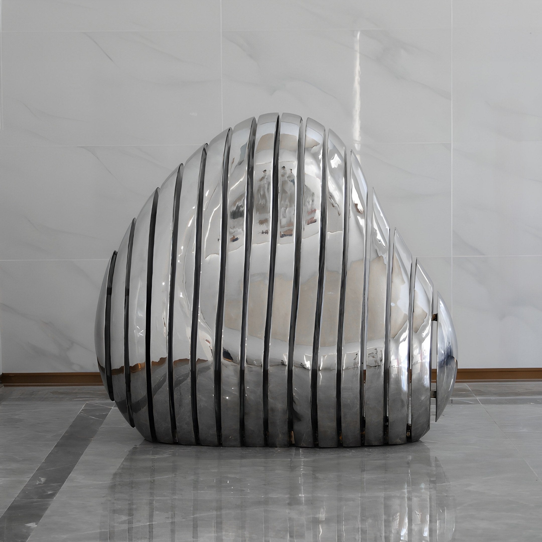 The Ripple Silver Organic Outdoor Steel Sculpture by Giant Sculptures features shiny, reflective metal strips forming a smooth, rounded design. It stands in an indoor space with marble-tiled flooring and a light-colored textured wall in the backdrop.