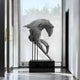 The Horse Silver Steel Abstract Sculpture (75/250cm) by Giant Sculptures, resembling a horses head, elegantly sits on a black pedestal in the modern hallway. Sunlight pours through a large window behind it, casting soft shadows on the floor.