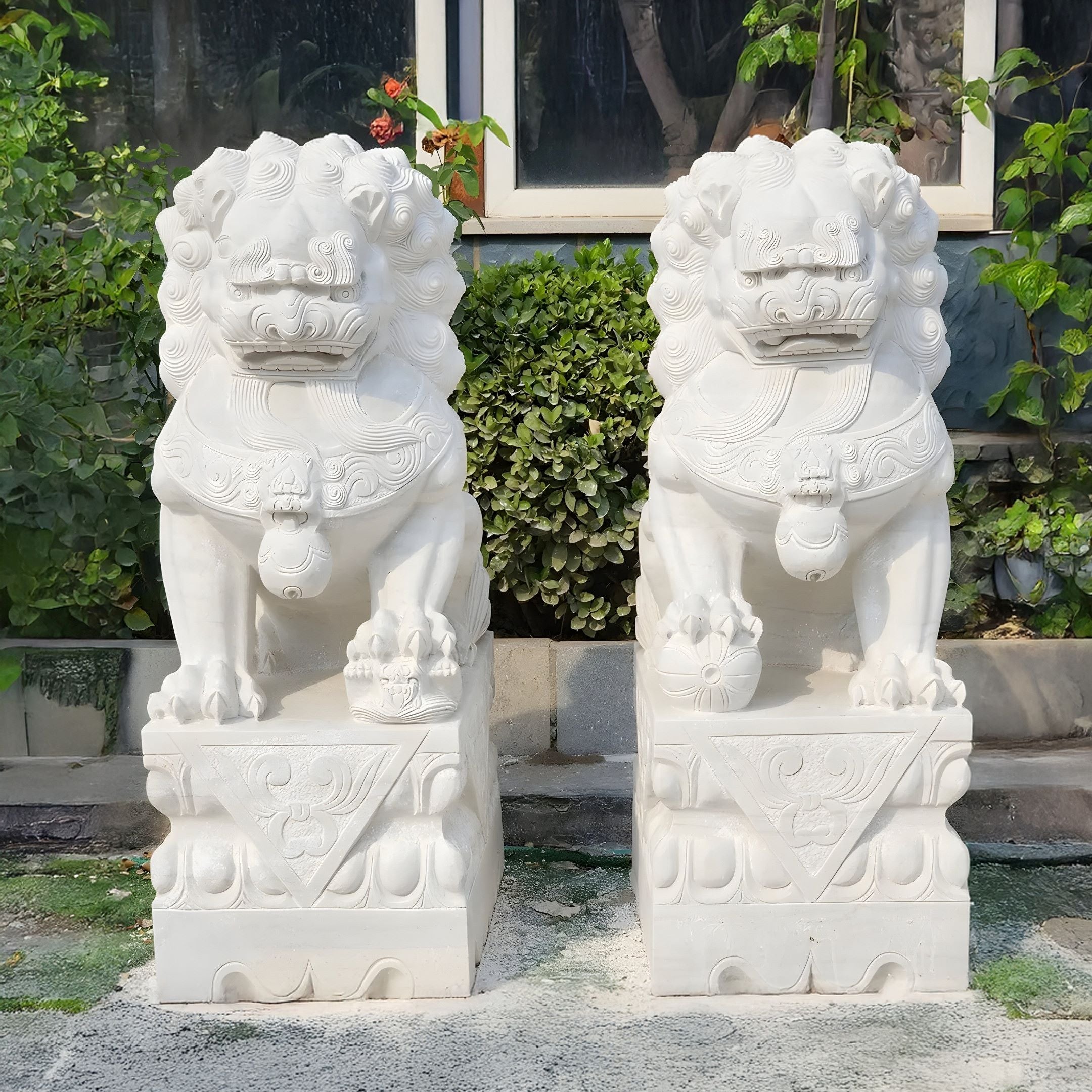 Two Chinese Guardian Lion IV Marble Sculptures by Giant Sculptures stand side by side on patterned square pedestals, displayed amidst greenery and against a glass window.