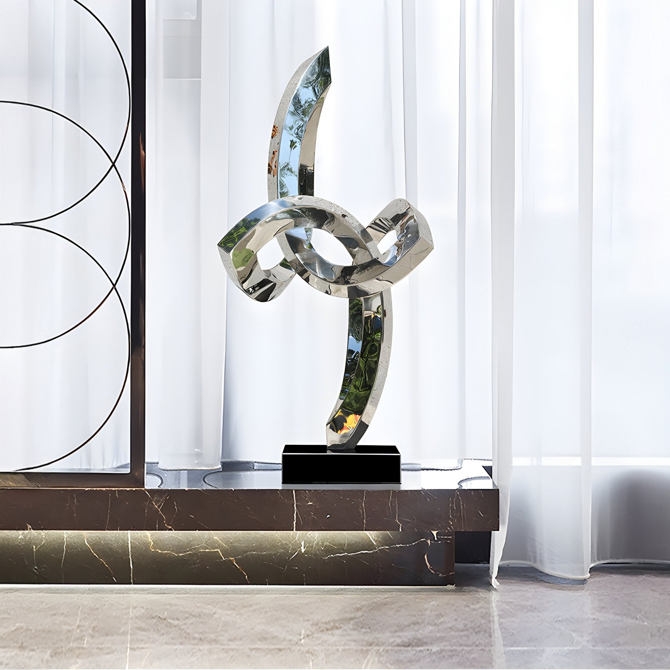 The Infinity Twist Abstract Steel Sculpture - 96cm by Giant Sculptures, featuring interlocking reflective loops on a black base, stands elegantly against a white curtain. Sunlight filters through the window, casting a soft glow on the polished marble floor and enhancing its contemporary design.