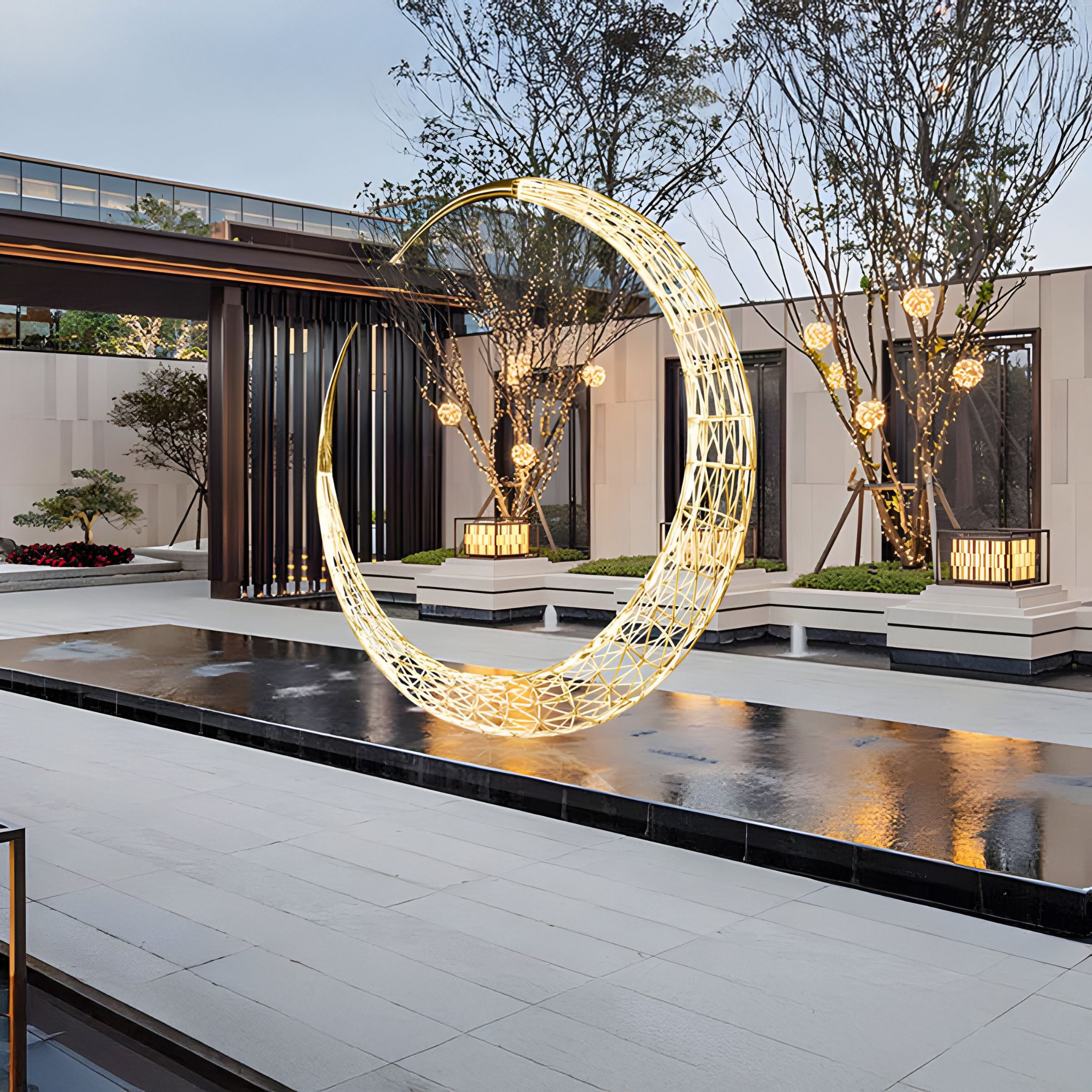 A modern courtyard features the Crescent Gold LED Wireframe Outdoor Steel Sculpture by Giant Sculptures, illuminating a tranquil reflecting pool. Surrounded by manicured trees and shrubs in raised planters, warm lighting accentuates this minimalist architectural space.