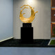 The Timeless Wave Gold Organic Steel Sculpture (117cm) by Giant Sculptures is a golden abstract piece resembling a swirling wave, mounted on a black pedestal in a sleek modern space with white walls and accented by the gentle presence of a green plant.