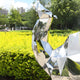 The Camel Geometric Steel Outdoor Sculpture from Giant Sculptures, standing 161/210cm tall, is a shiny art piece resembling an abstract bird in the park. Its framed by vibrant green foliage and yellow flowers, with tall trees and city buildings set against a clear blue sky.