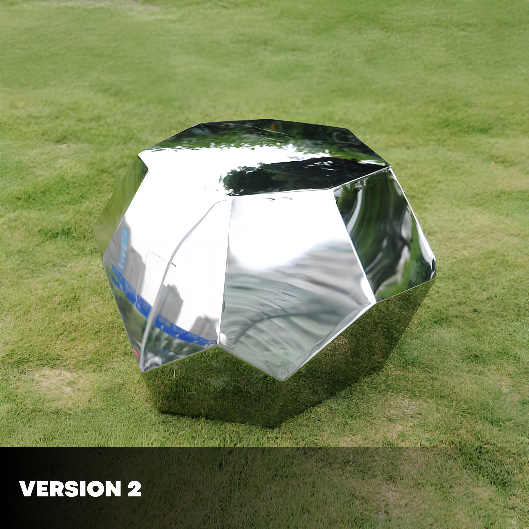 The Tetra Geometric Steel Rock Sculpture by Giant Sculptures rests on green grass, its mirrored facets reflecting the surroundings with contemporary elegance. The lower left corner displays the text: VERSION 2.