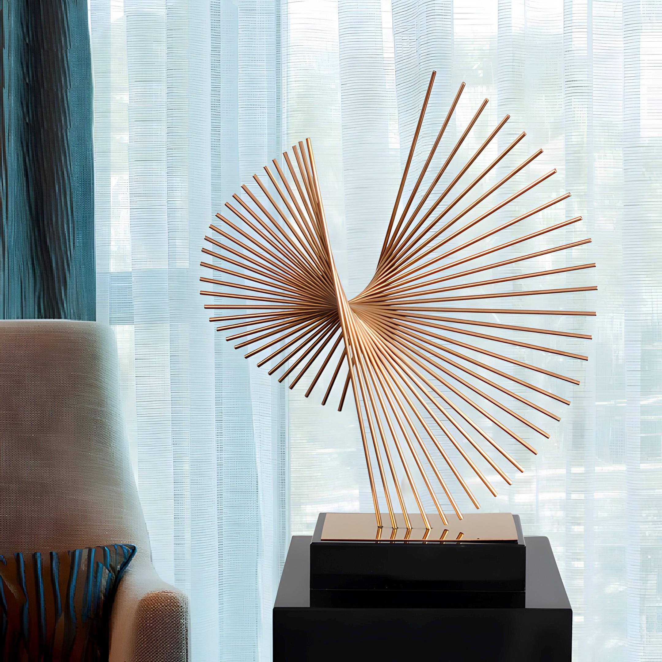 The Axis Gold Abstract Steel Sculpture by Giant Sculptures, featuring intersecting gold rods on a dark pedestal, is beautifully showcased with sheer curtains and a beige chair with a striped pillow in the background—capturing the essence of luxury garden art.