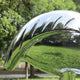 The Crane Contemporary Outdoor Steel Sculpture from Giant Sculptures is a curved leaf with notched edges made of stainless steel. Its reflective surface mirrors nature, creating an abstract interplay of light and lush greenery in this striking 98/158cm sculpture.