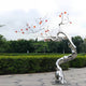 A shiny silver abstract sculpture standing outdoors, reflecting its surroundings on the shiny surface of the tree trunk.