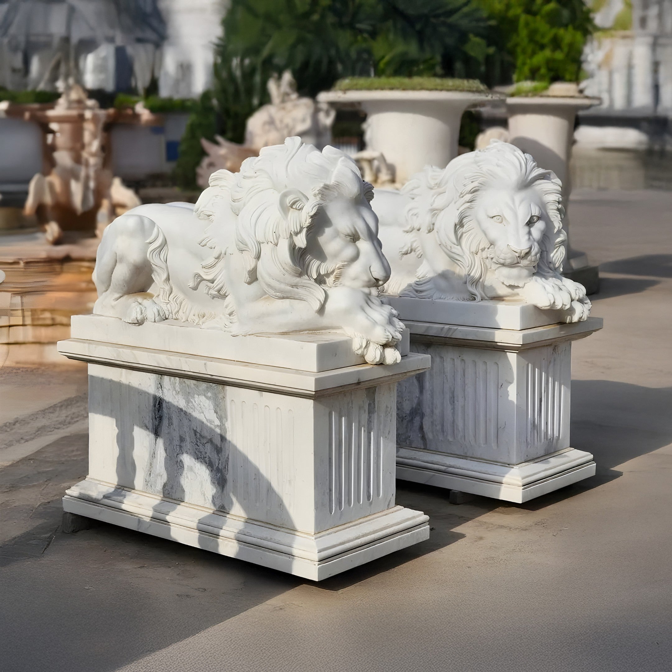 The Giant Lying Lion Marble Outdoor Sculptures from Giant Sculptures rest gracefully on rectangular pedestals, basking in sunlight. Surrounded by lush greenery and additional sculptures, these exquisite pieces enhance the refined aesthetic of the garden setting.