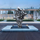 In a modern outdoor courtyard, the Molten Silver Organic Steel Sculpture by Giant Sculptures stands on a pedestal, accompanied by a sleek building with large windows and trees under a clear sky. This 155cm stainless steel masterpiece gleams brilliantly amidst its serene surroundings.