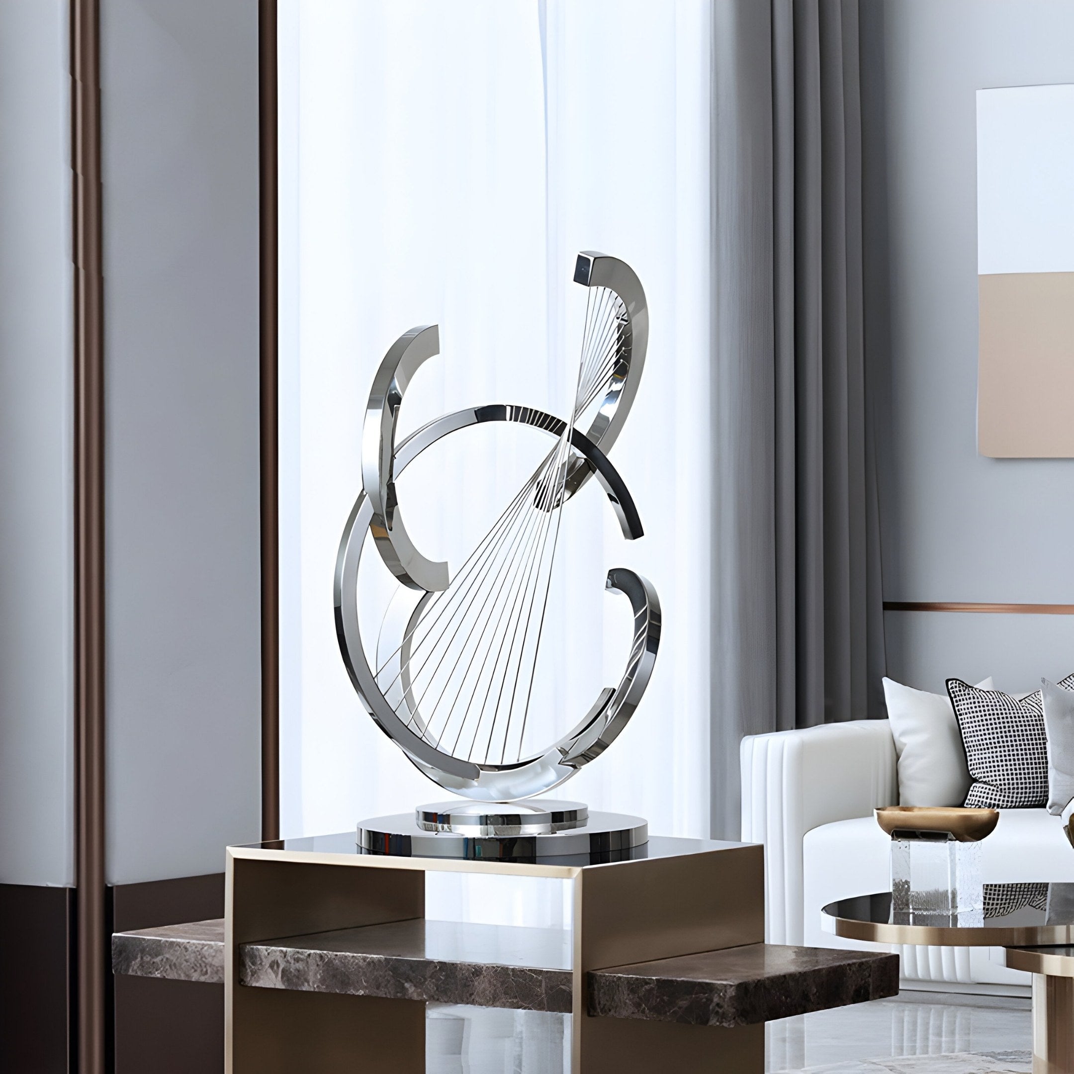 The Orbital Strings Abstract Steel Sculpture - 80cm by Giant Sculptures, a modern piece with intertwined metallic curves, elegantly adorns a pedestal in a stylish living room with a white sofa, neutral cushions, and large windows that highlight its minimalist design.